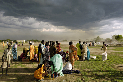Alcuni profughi raggruppati in un campo in Sudan ricevono razioni di cibo; sullo sfondo il cielo scuro preannuncia l'arrivo di una tempesta.