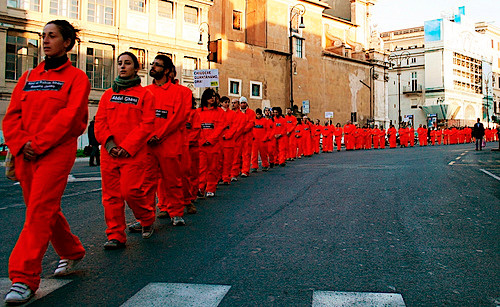 Una fila di persone sfila per la strada con le tute arancioni simili a quelle che indossano i detenuti a Guantanamo