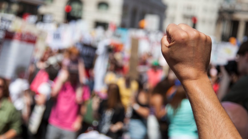 L'immagina mostra sullo sfondo un gruppo di persone che manifestano in strada e in primo piano un braccio con il pugno alzato.