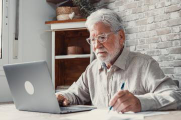 Older person in front of a laptop while he writes with a pen on a paper sheet 