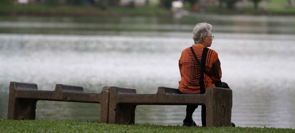 An elderly woman sits alone on a bench near a pond in Thailand