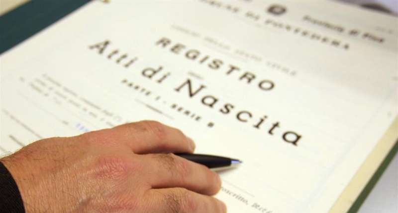 Immagine di una mano con una penna fra le dita e in basso un atto di nascita con scritto "Registro Atto di Nascita"