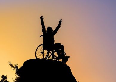 Una persona in sedia a rotelle si erge con le braccia alzate in cima a una montagna