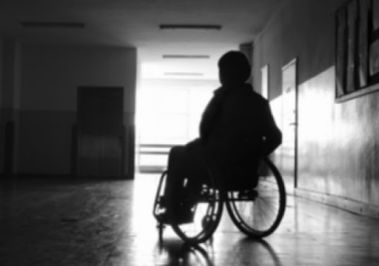 L'immagine rappresenta una persona con disabilità in carrozzina, in un corridioio. L'immagine è molto buia e non è possible riconoscere l'identità della persona fotografata, né altre sue caratteristiche.