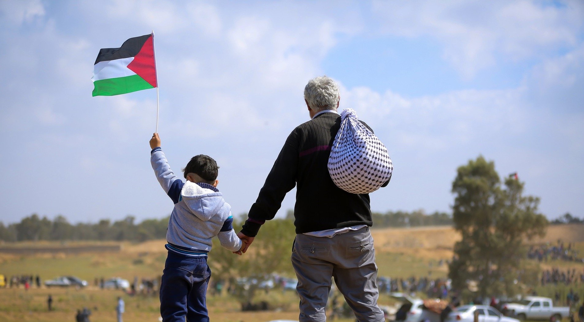 Un bambino per mano al nonno sono in piedi su una macchina e guardano l'orizzonte. Il bambino sventola una bandiera palestinese e il nonno ha una kefiah sulle spalle