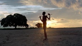 Foto in controluce di un giovane dalla figura esile che tiene sulle spalle un bastone, in un campo di Bahr El Ghazal, Sudan. 