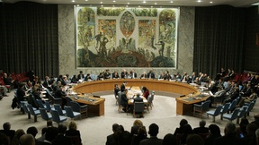 A UN Security Council session. The participants are seated around a circular table, in the center of which is positioned another, rectangular table. 