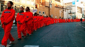 Una fila di persone sfila per la strada con le tute arancioni simili a quelle che indossano i detenuti a Guantanamo