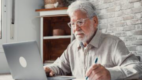 Una persona anziana davanti ad un computer portatile mentre scrive con una penna su un foglio di carta 