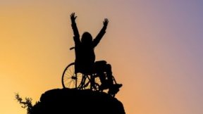 Una persona in sedia a rotelle si erge con le braccia alzate in cima a una montagna