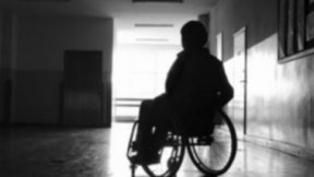 L'immagine rappresenta una persona con disabilità in carrozzina, in un corridioio. L'immagine è molto buia e non è possible riconoscere l'identità della persona fotografata, né altre sue caratteristiche.
