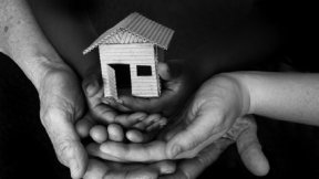 Black and white photo - an adult and child's hands holding a small house made of cardboard