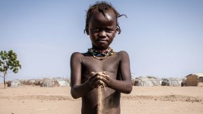  African child holds desert's sand in his hands. A village in the background. 