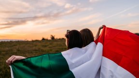 Due ragazze abbracciate con la bandiera italiana sulle spalle guardano un campo
