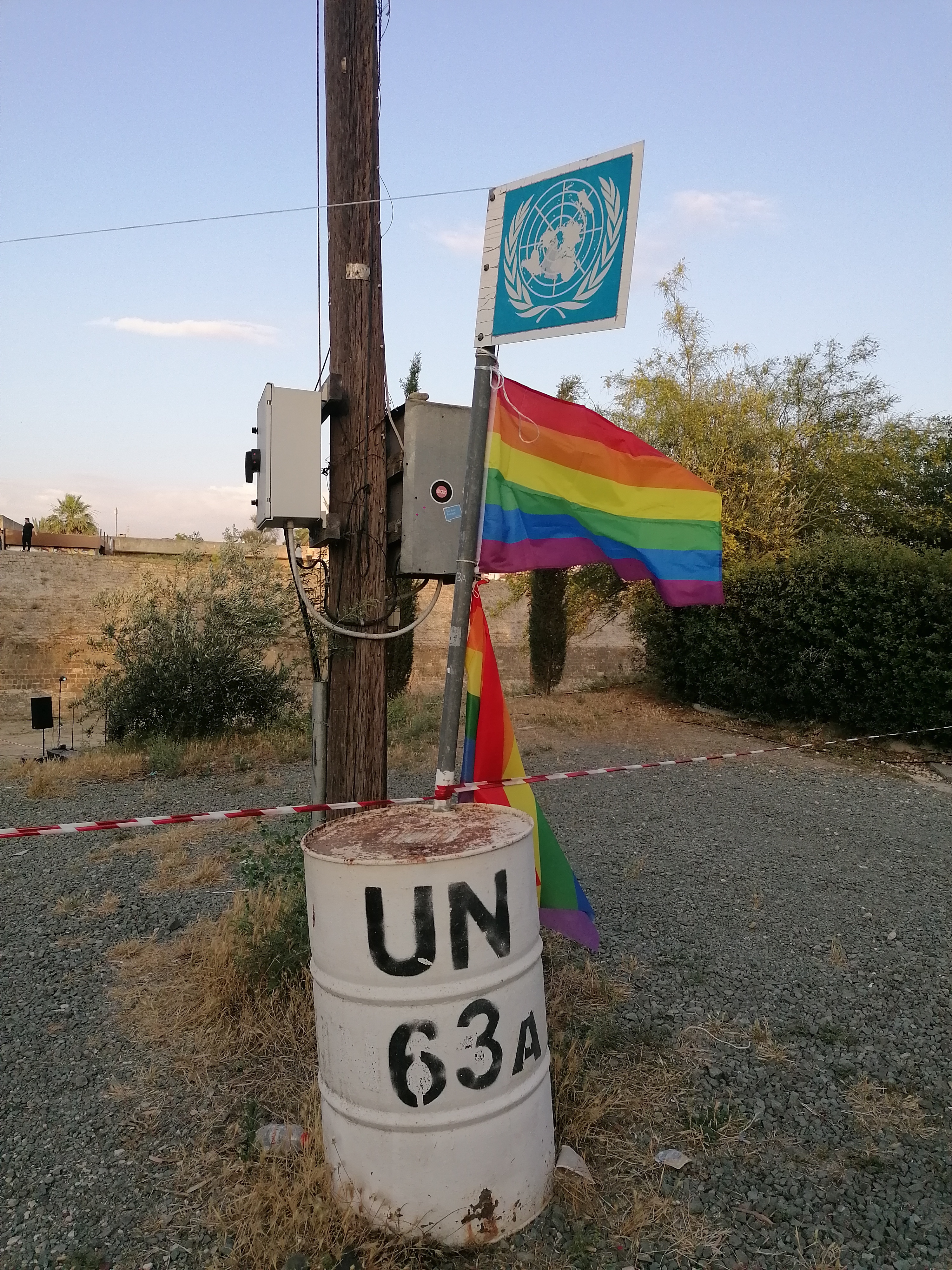 Foto di un barile che delimita la zona cuscinetto di Cipro. Vicino l'egida delle Nazioni Unite sono appese due bandiere arcobaleno.